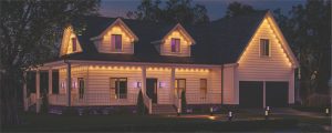 An outdoor porch with wooden flooring featuring a White couch and ottoman, Orange pillows and blankets. Green bushes in the background, with a Hue Go Portable Table Lamp set up on the ottoman. With dim natural light in the background of the setting, Hue outdoor lights provide soft tones of Purple.