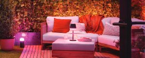 Une véranda extérieure au plancher de bois comprend un canapé et un repose-pieds blancs, agrémentés de coussins et de couvertures orange. Des arbustes verts se trouvent à l’arrière-plan, tandis qu’une lampe de table portative Hue Go est posée sur le repose-pieds. Dans la douce lumière naturelle du crépuscule, les luminaires extérieurs Hue diffusent de délicates teintes violettes.