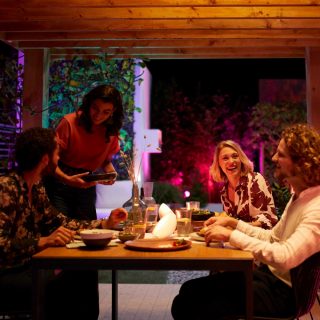 Un groupe d'amis prend un repas dehors, sur un patio décoré avec des lampes connectées colorées.