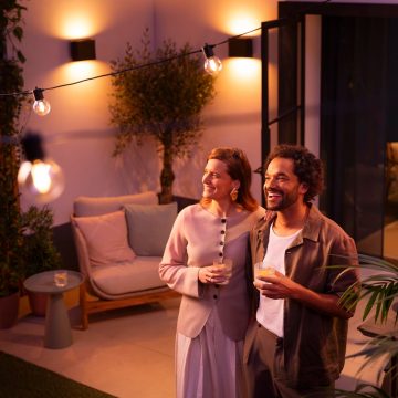 To personer står på en terrasse under Philips Hue-lyslenker og vegglamper, med drikke i hånden i et varmt opplyst uteområde.