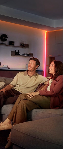 A couple sit on a sofa in a living room with corners and ceiling lines lit with direct light from an OmniGlow strip light. 