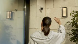 Eine Frau an der Haustür winkt in eine Hue Secure Überwachungskamera, die an einer Wand befestigt ist. 