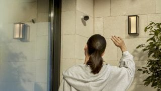A woman at a front door waving to a Hue Secure security camera mounted on the wall. 