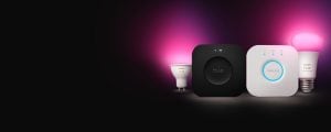 A Hue Bridge Pro smart home hub and a Philips Hue Bridge smart home hub shown with smart bulbs glowing in pink light, all included in the Philips Hue create your own starter kit promotion. 