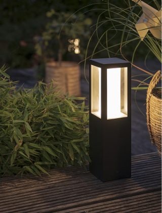 A black pedestal smart light placed in a garden and glowing in warm white light.