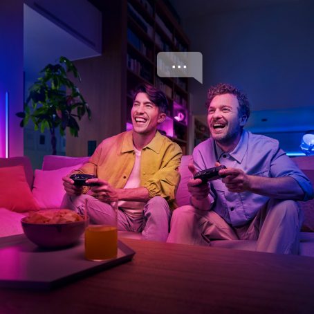 Two men on couch laughing while playing a video game using their voices to control the Hue sync box.
