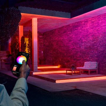 A person uses the Hue app on his smartphone to control concealed Flux outdoor strip lights on a patio glowing in pink and orange light. 