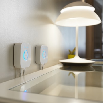 Two white smart lighting hubs mounted on a wall above a glass-topped desk, illuminated by a modern white table lamp.