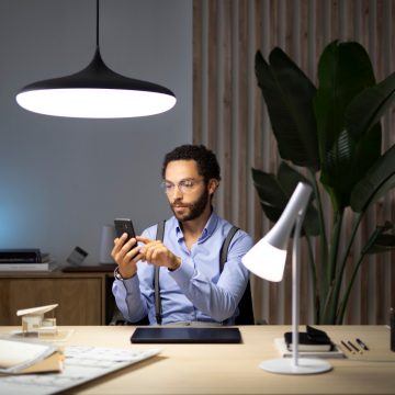 Person using a smartphone at a desk in a modern office, illuminated by pendant and desk smart lights, with plants in the background.