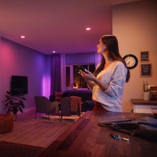 Person adjusting smart ambient lighting in a modern living room using a mobile device, with colored lights illuminating seating area.