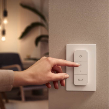 Finger pressing the brightness increase button on a white smart lighting wall switch labeled ""hue"" mounted on a light-colored wall.