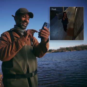 A person outdoors by a lake uses a smartphone to check a home security camera feed showing a visitor at a well-lit front porch.