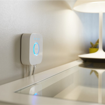 A square white smart lighting hub is mounted on a wall above a desk, with indicator lights illuminated and cables connected.