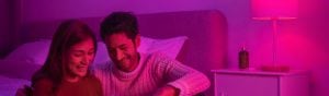 Couple sitting together on a bed in a bedroom with smart lamp casting pink light over the bedside table and headboard.
