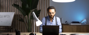 Person working at a desk with bright task lighting, laptop, desk lamp, and architectural plans in a modern study setting.