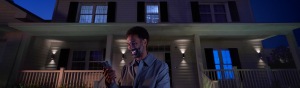 Person standing on a porch at night using a smartphone, with outdoor wall lights illuminating the house entrance.