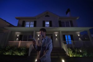 Person with a smartphone for managing a home automation system in front of a house at night with smart outdoor lighting illuminating the porch and garden. 