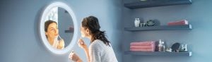 Person applying makeup at a vanity mirror, illuminated by a circular mirror light in a modern bathroom with shelves and towels.
