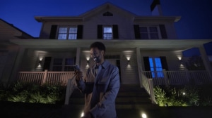 Person standing in front of a house at night using a smartphone, with security lighting outdoor illuminating the entrance.