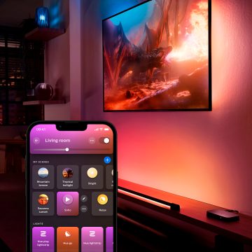 A smart light bar placed on a cabinet projects colored light onto a wall behind a TV, controlled by a mobile app shown in the foreground.