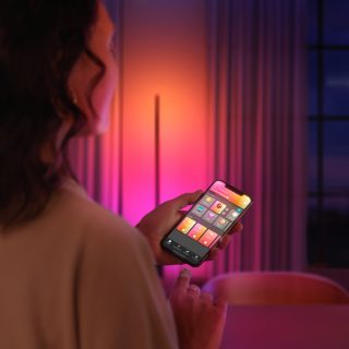 Person adjusting smart lighting settings on a mobile app in a living room with colorful ambient light in the background.