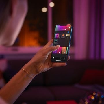Person using a smartphone app to adjust colorful smart lighting in a living room setting with purple and pink ambient lights.