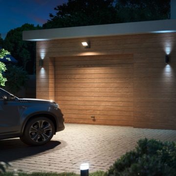 Wall-mounted and ground smart lighting fixtures illuminate a wooden garage door and driveway, enhancing visibility at night.