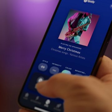 A hand holds a smartphone displaying a music app with ""Merry Christmas"" by Various Artists, showing style, intensity, color, brightness buttons.