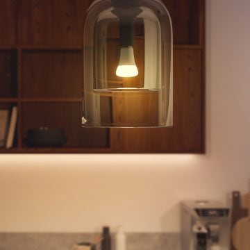 A smart light bulb emitting white light is installed in a glass pendant fixture above a kitchen counter with wooden shelves.