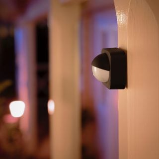 Black and white motion sensor mounted on an exterior wall, positioned to monitor entryway with outdoor smart lights in the background.