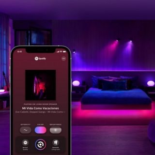 A bedroom with smart lights displaying purple and pink tones, syncing with music shown on a smartphone Spotify interface.