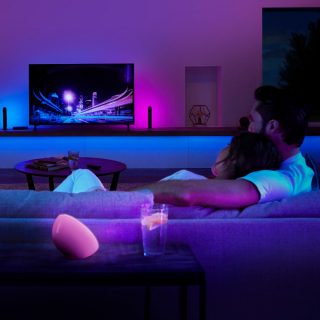 Two people sit on a sofa watching TV in a living room with colorful smart lighting synced to the television screen.