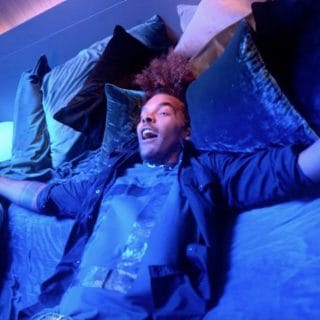 Person relaxing on a bed surrounded by pillows, illuminated by vibrant blue and purple smart lighting in a bedroom setting.