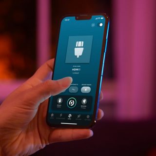 A hand holds a phone displaying the Hue Sync app, showing HDMI 1 linked, intensity and brightness sliders, and sync controls.