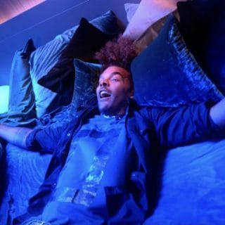 Person relaxing on a bed surrounded by pillows, illuminated by vibrant blue and purple smart lighting in a bedroom setting.