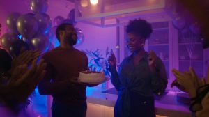 Group celebrating a birthday in a kitchen with colorful ambient lighting, balloons, and a cake being presented.