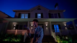 A person using a smartphone stands outside a house at night, with smart outdoor lighting illuminating the porch and garden.