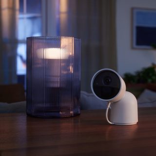 White indoor security camera positioned on a wooden table beside a translucent lamp, both devices in clear focus.