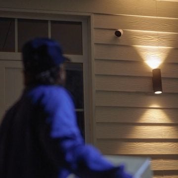 A person approaches a home entrance at night, illuminated by outdoor wall lighting and monitored by a security camera.