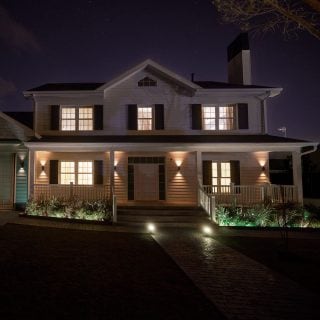 A white house at night with smart security cameras and exterior lighting mounted above the porch, illuminating the entrance and pathway.