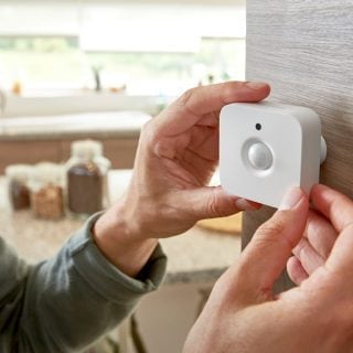 Hands mounting a white motion sensor on a wooden wall, likely for smart lighting or security automation purposes.