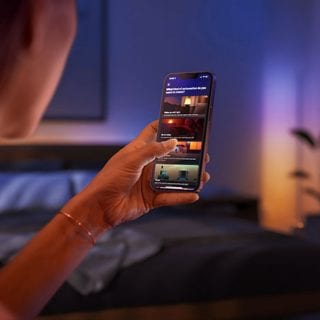 Person adjusting smart lighting automations on a smartphone in a bedroom, with colored lights illuminating the background.