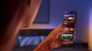 Person using a smartphone to set lighting automation, with text options like ""Wake up with light"" and ""Go to sleep"" visible in a bedroom.