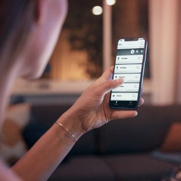Person adjusting smart lighting settings on a smartphone in a living room with warm light illuminating the background.