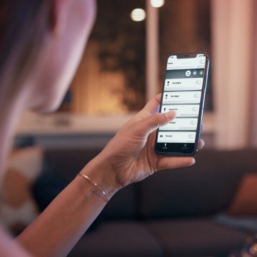 Person adjusting smart lighting settings on a smartphone in a living room with warm light illuminating the background.