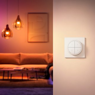 Wall-mounted white smart lighting dimmer switch in focus, positioned beside a living area with pendant lights in the background.