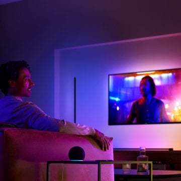 A person sits on a sofa in a living room watching TV, with colorful ambient lighting illuminating the space.