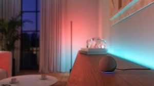 A spherical smart speaker with a blue light ring sits on a wooden surface, illuminated by colored smart lighting on the wall.