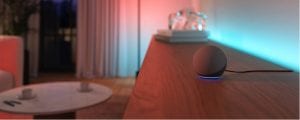 A spherical smart speaker with a blue light ring sits on a wooden surface, with colored lighting illuminating the wall behind.