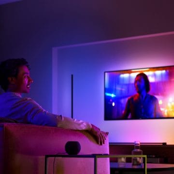 Person seated on a sofa in a living room watching TV, with colored ambient lighting illuminating the wall behind the screen.